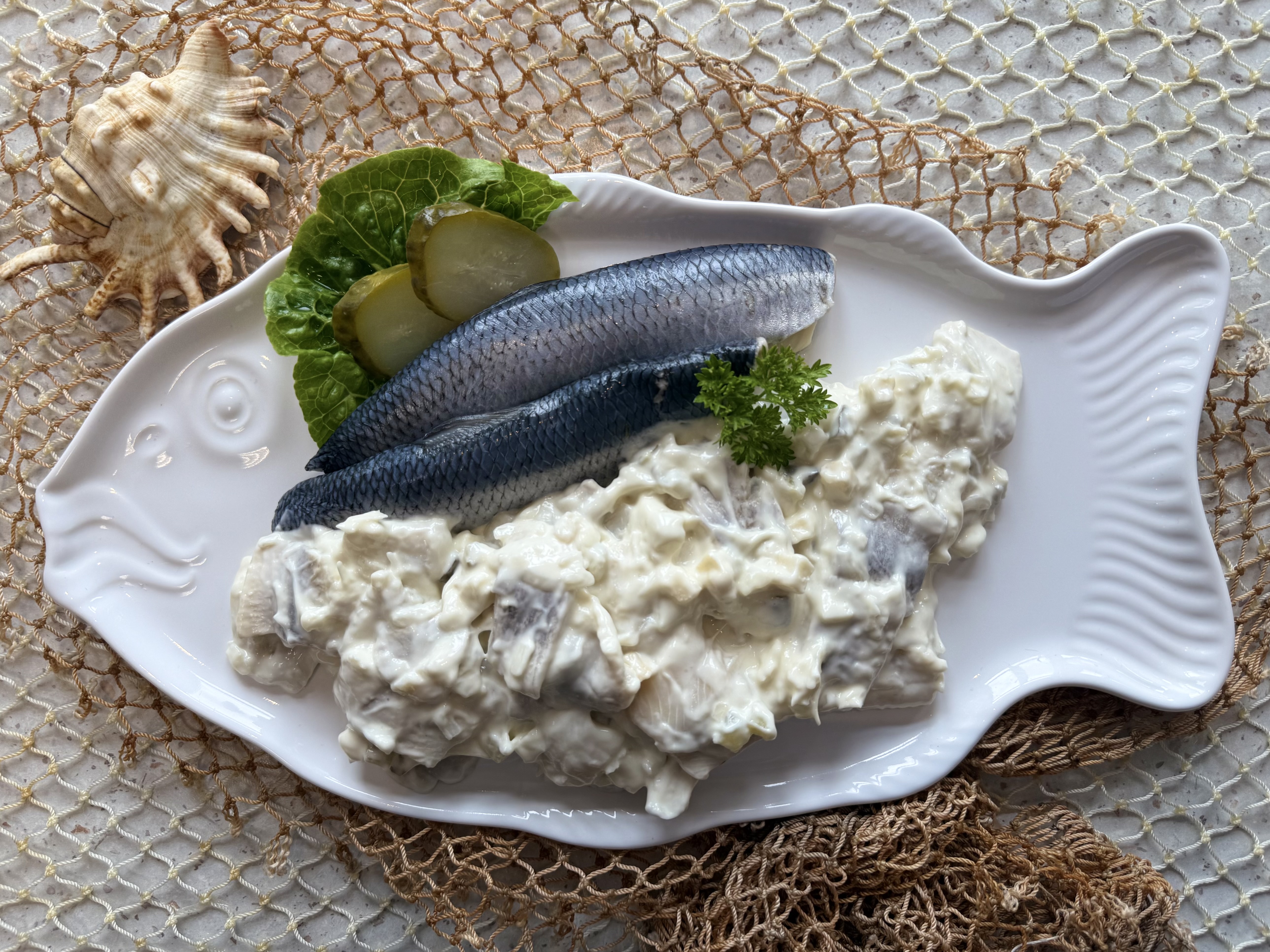 Weißer Heringssalat mit Heringfilets & Gurkenscheiben auf Salatblatt auf weißem Teller.