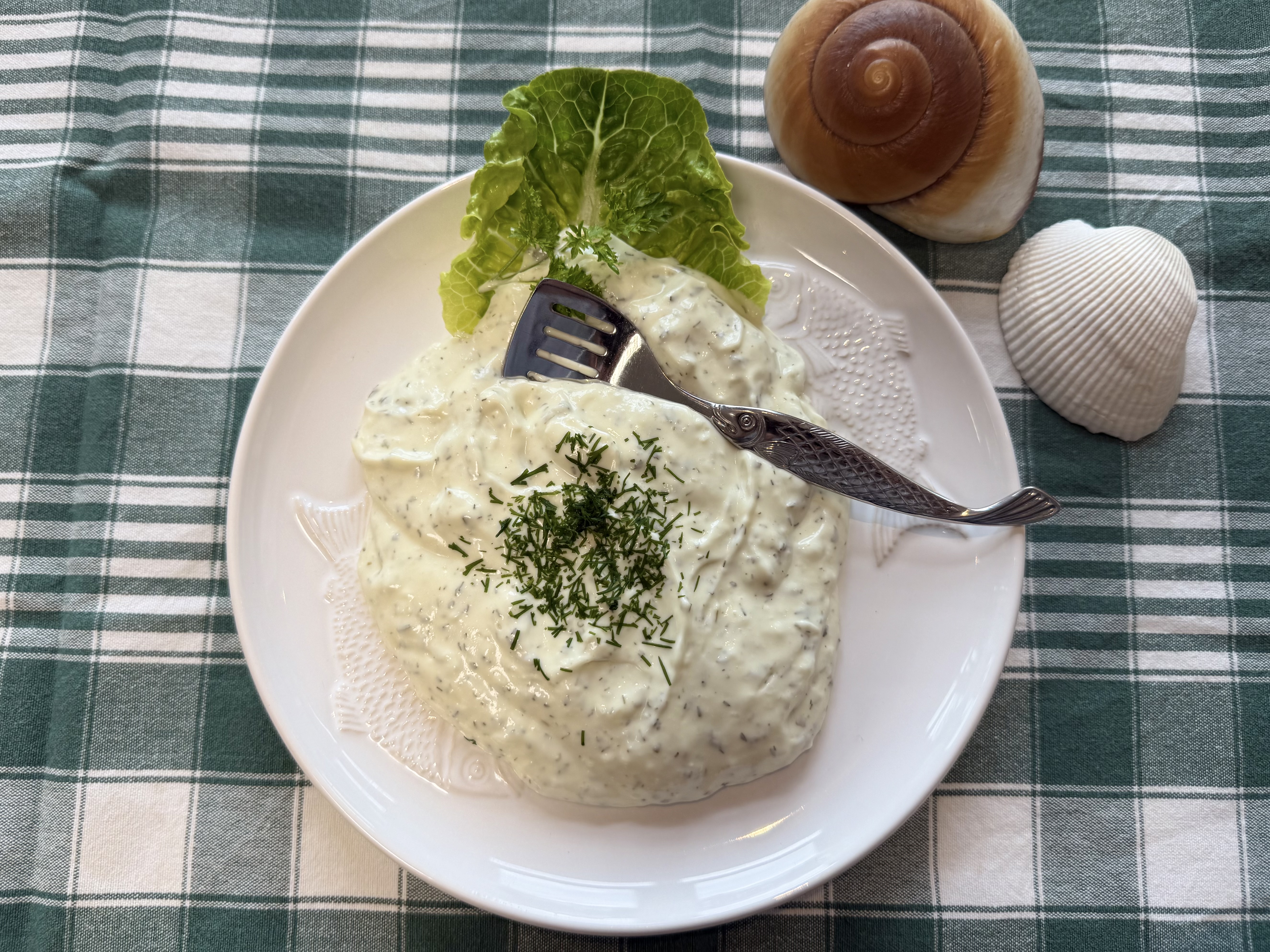Cremige Kräuter-Remoulade auf Teller, garniert mit Dill; Salatblatt und Meeres-Deko im Hintergrund.